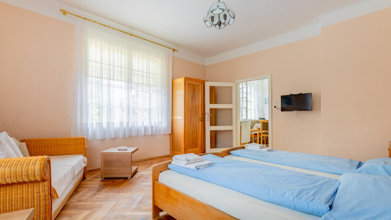 Ein gemütliches Schlafzimmer mit zwei Einzelbetten, einem Sofa, einem Schrank und einem kleinen Tisch. Ein Fenster mit Vorhängen lässt Tageslicht herein.