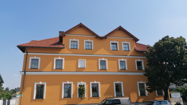Frühstückspension Csenar, © Wiener Alpen Gelbes Gebäude mit roten Dachziegeln und mehreren Fenstern, davor geparkte Autos und ein Baum.