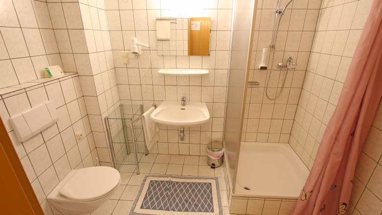 Kleines Badezimmer mit Dusche, Waschbecken und Toilette, gefliest in Weiß.