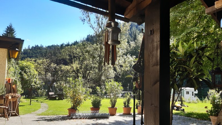 Ein sonniger Innenhof mit Rasen, Bäumen und Topfpflanzen. Im Vordergrund eine überdachte Terrasse mit einer Laterne und Gartenmöbeln.