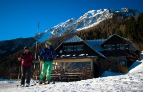 Zwei Skifahrer vor der Edelweisshütte am Schneeberg mit schneebedecktem Berg im Hintergrund.