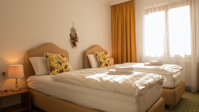 Ein gemütliches Doppelzimmer mit zwei Einzelbetten, gelben Vorhängen und einem Traumfänger an der Wand.