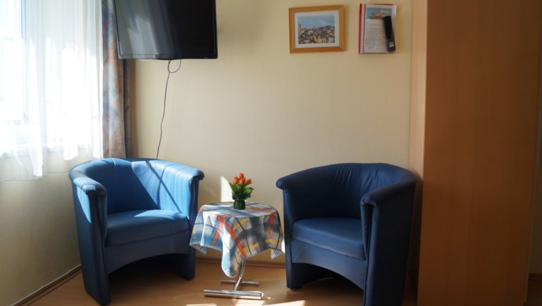 Zwei blaue Sessel mit einem kleinen Tisch dazwischen, darauf eine Vase mit Blumen. Ein Fernseher hängt an der Wand, daneben zwei Bilder.