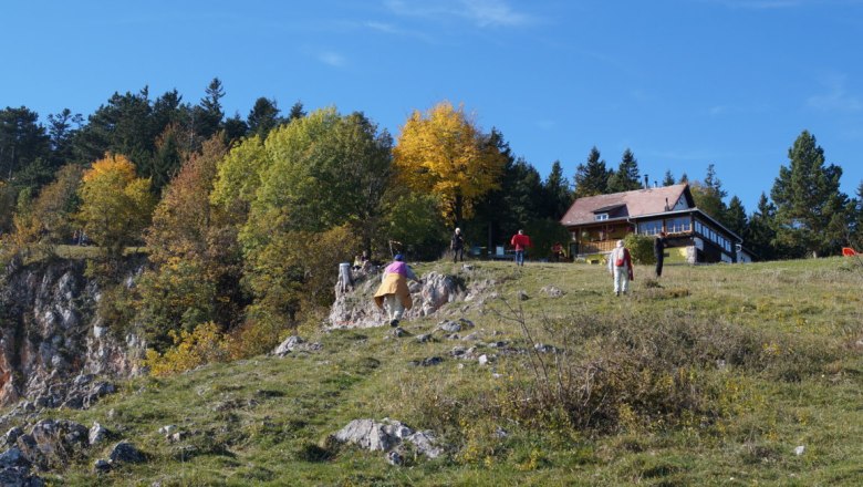 Menschen wandern auf einem grasbewachsenen Hügel mit einer Berghütte im Hintergrund.