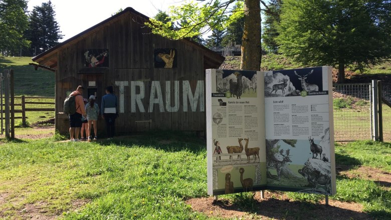 Eine Familie steht vor einer Holzh&uuml;tte mit der Aufschrift 'TRAUM'. Daneben eine Infotafel &uuml;ber Tiere in einem gr&uuml;nen Park.