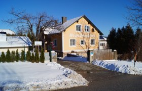 Ein gelbes Haus mit der Aufschrift 'Haus Bergblick' im Schnee, umgeben von Bäumen und einem blauen Himmel.