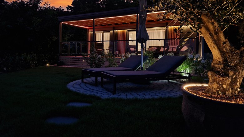 Außenansicht eines Chalets bei Sonnenuntergang mit Liegestühlen und beleuchtetem Baum im Vordergrund.