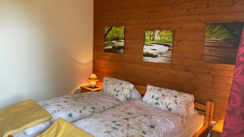 Gem&uuml;tliches Doppelzimmer mit Holzw&auml;nden, einem Doppelbett mit Blumenbettw&auml;sche und gelben Decken, sowie einer Nachttischlampe.