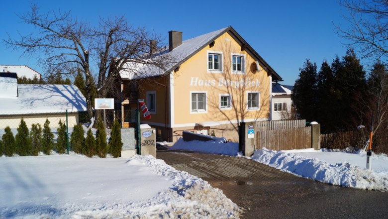 Ein gelbes Haus mit der Aufschrift 'Haus Bergblick' im Schnee, umgeben von Bäumen und einem blauen Himmel.