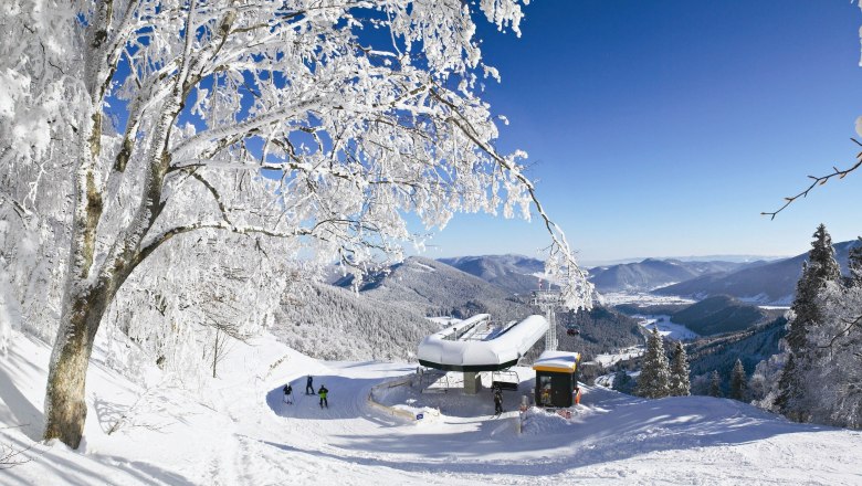 Winterlandschaft mit Sesselliftstation, verschneiten B&auml;umen und Bergen im Hintergrund.