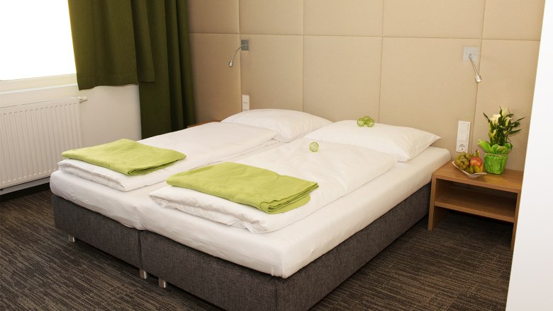 Gemütliches Hotelzimmer mit Doppelbett, grünen Handtüchern und Nachttisch mit Blumen und Obst.