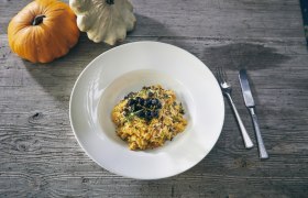 Ein Teller K&uuml;rbisrisotto mit Kr&auml;utern und Beeren, daneben zwei K&uuml;rbisse auf einem Holztisch.