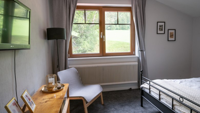 Schlafzimmer mit grauem Teppich, Bett, Sessel, Fernseher und Fenster mit Blick ins Gr&uuml;ne.