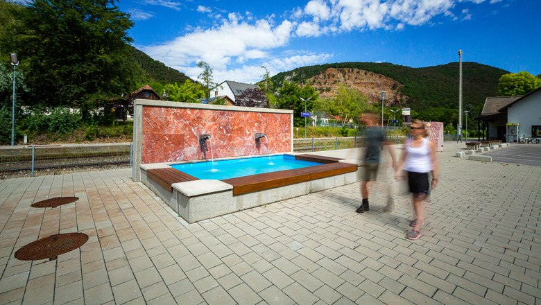 Moderner Brunnen auf einem Platz mit zwei unscharfen Personen im Vordergrund, umgeben von B&auml;umen und H&uuml;geln.
