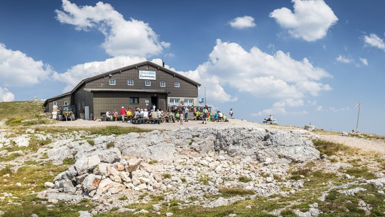Fischerhütte auf dem Schneeberg mit Wanderern und blauem Himmel.