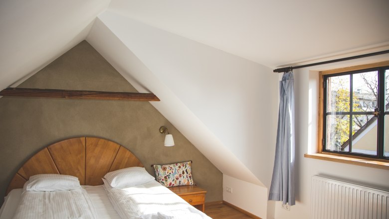 Gemütliches Schlafzimmer mit Dachschräge, Doppelbett, Holzdetails und Fenster mit Vorhang.