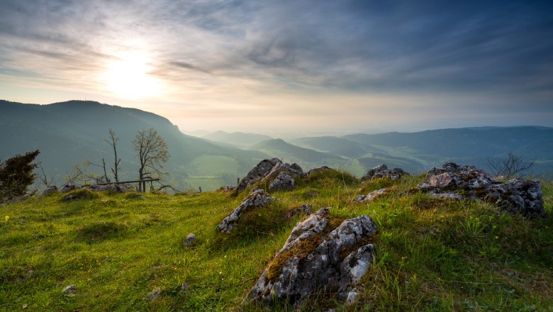 Panoramablick von einem H&uuml;gel mit Felsen und Gras im Vordergrund, Bergen und einem bew&ouml;lkten Himmel im Hintergrund.