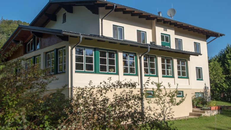 Ein zweistöckiges Haus mit vielen Fenstern und einem Satteldach, umgeben von Bäumen und Sträuchern, unter einem klaren blauen Himmel.
