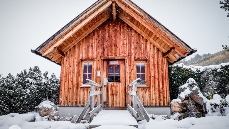 Eine kleine Holzhütte im Schnee mit einem steilen Dach und zwei Fenstern.