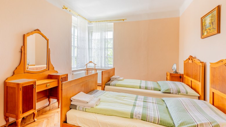 Ein traditionelles Schlafzimmer mit zwei Einzelbetten, einem Schminktisch mit Spiegel und einem Fenster mit weißen Vorhängen.