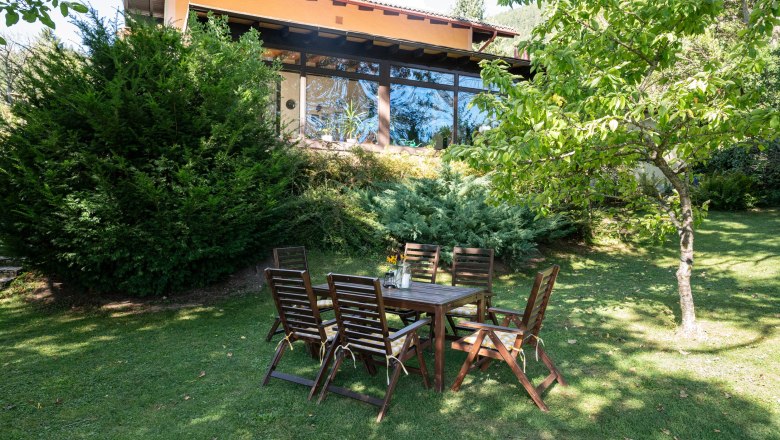 Ein Garten mit Holztisch und St&uuml;hlen vor einem Ferienhaus mit gro&szlig;en Fenstern und umgeben von B&auml;umen und Str&auml;uchern.