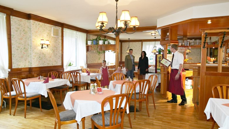 Ein rustikales Restaurant mit Holzmöbeln und Tischdecken. Ein Kellner begrüßt eine Familie am Eingang.