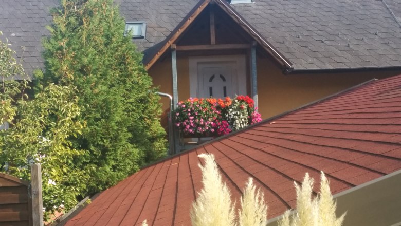 Hausdach mit rotem Schindeldach, Balkon mit Blumen, Baum im Hintergrund.