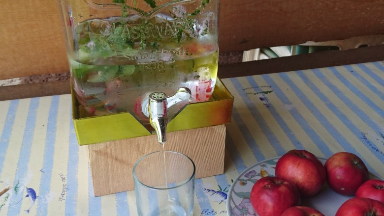 Ein Glasbehälter mit Wasser und Kräutern, daneben ein Glas und ein Teller mit roten Äpfeln auf einem gestreiften Tisch.