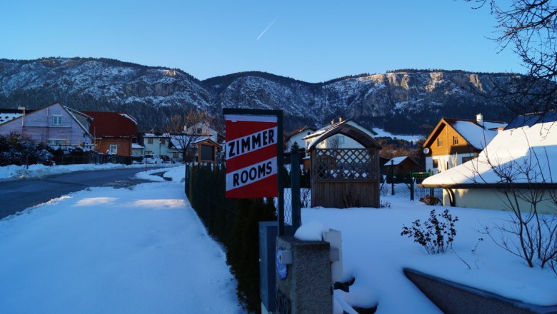 Winterliche Landschaft mit schneebedeckten Häusern und Bergen im Hintergrund. Ein Schild mit der Aufschrift 'Zimmer Rooms' ist sichtbar.