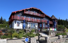 Ein großes, traditionelles Haus mit rotem Holz und weißen Wänden, umgeben von einem Garten und blauen Sonnenschirmen.