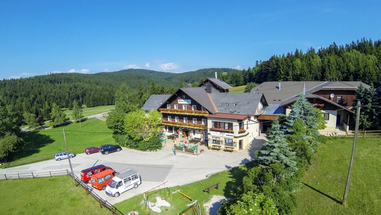 Alpengasthof Postl in einer grünen, bewaldeten Landschaft mit mehreren geparkten Autos und einem Spielplatz im Vordergrund.