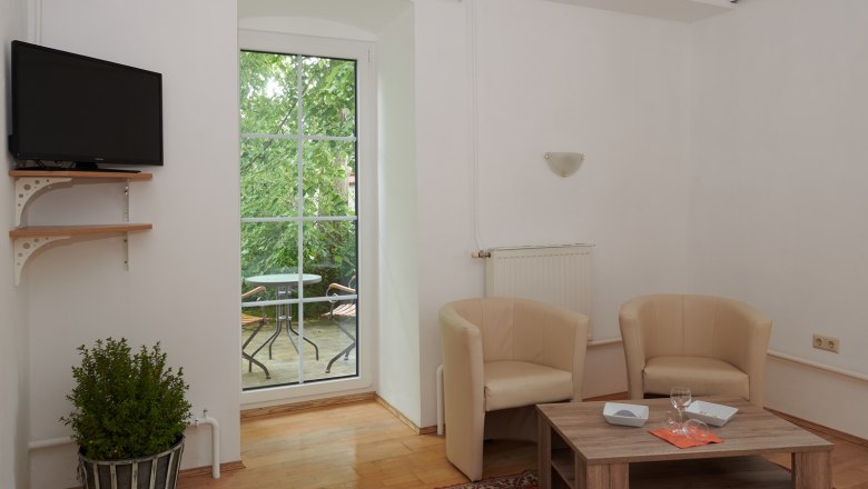 Wohnzimmer mit zwei beigen Sesseln, Holztisch, Fernseher und Balkon mit Tisch und Stühlen.