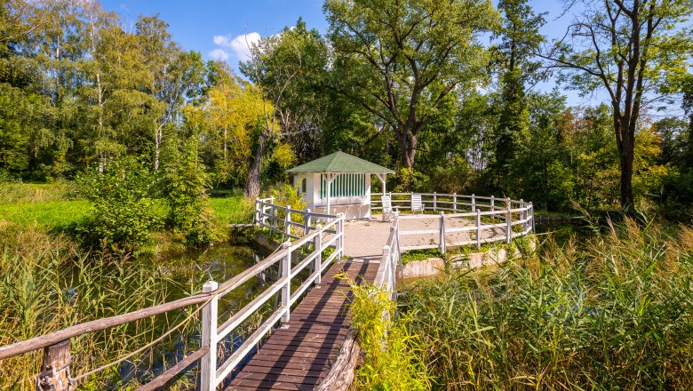 Ein Pavillon mit grünem Dach auf einer Plattform, umgeben von Bäumen und Schilf, mit einem Holzsteg als Zugang.