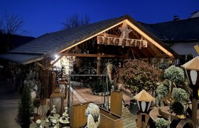 Ein dekorativ beleuchtetes Holzgeb&auml;ude mit der Aufschrift 'Deko Stadl' bei Nacht, umgeben von Pflanzen und Dekorationsartikeln.