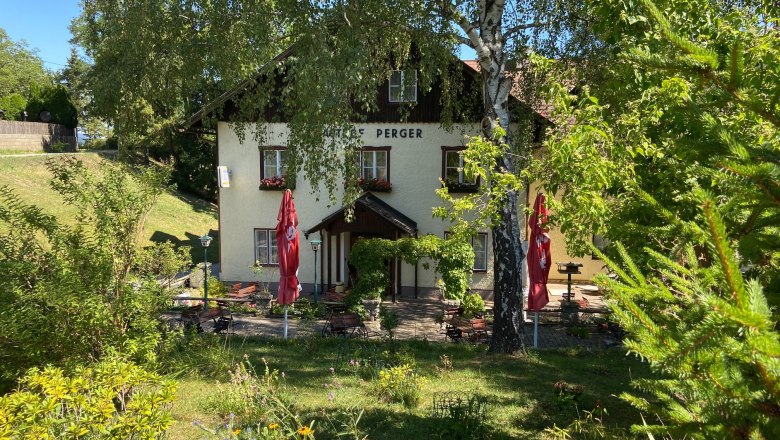 Ein Gasthaus mit Terrasse, umgeben von Bäumen und Pflanzen, bei sonnigem Wetter.