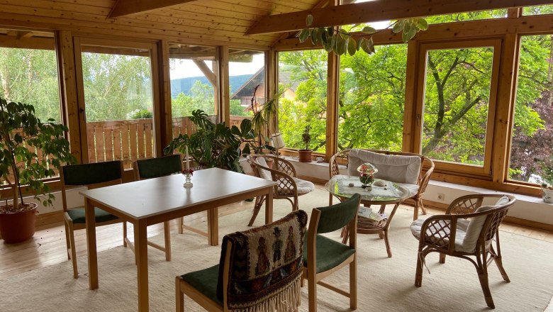 Ein heller Raum mit Holzm&ouml;beln, gro&szlig;en Fenstern und Pflanzen.