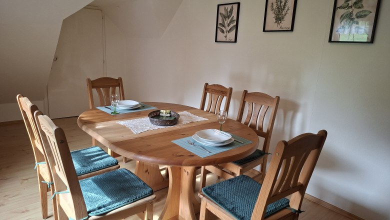 Essbereich mit Holztisch und sechs St&uuml;hlen, dekoriert mit blauen Sitzkissen. An der Wand h&auml;ngen drei botanische Bilder.