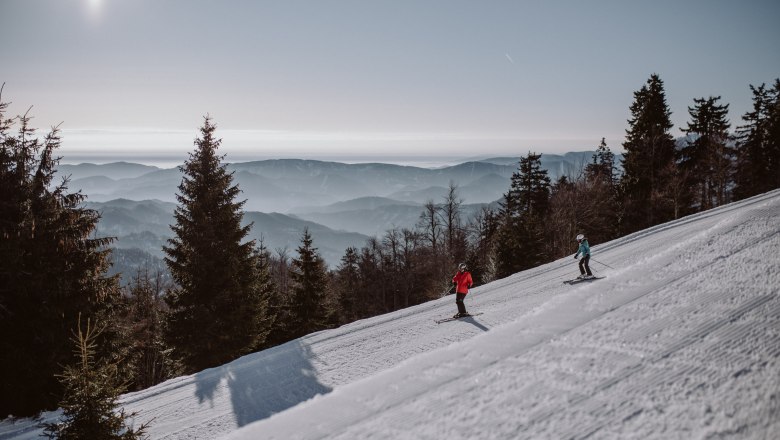 Zwei Skifahrer auf einer Piste im Schigebiet Unterberg mit Bergpanorama im Hintergrund.