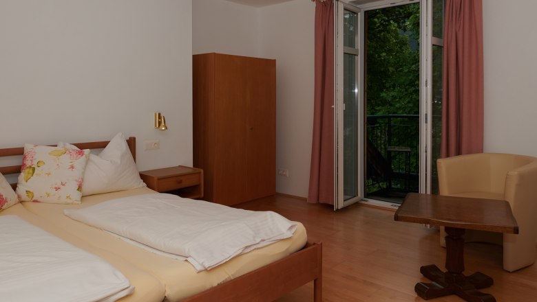 Schlafzimmer mit Doppelbett, Schrank, Tisch und Stuhl, Fenster mit Vorhängen.