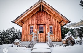 Eine kleine Holzh&uuml;tte im Schnee mit einem steilen Dach und zwei Fenstern.