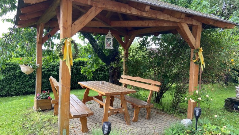 Holzlaube im Garten mit Tisch und B&auml;nken, umgeben von Pflanzen.