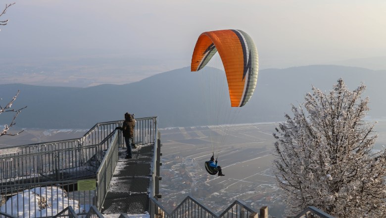 Ein Paraglider fliegt &uuml;ber eine verschneite Landschaft im Naturpark Hohe Wand, w&auml;hrend eine Person auf einer Aussichtsplattform steht.