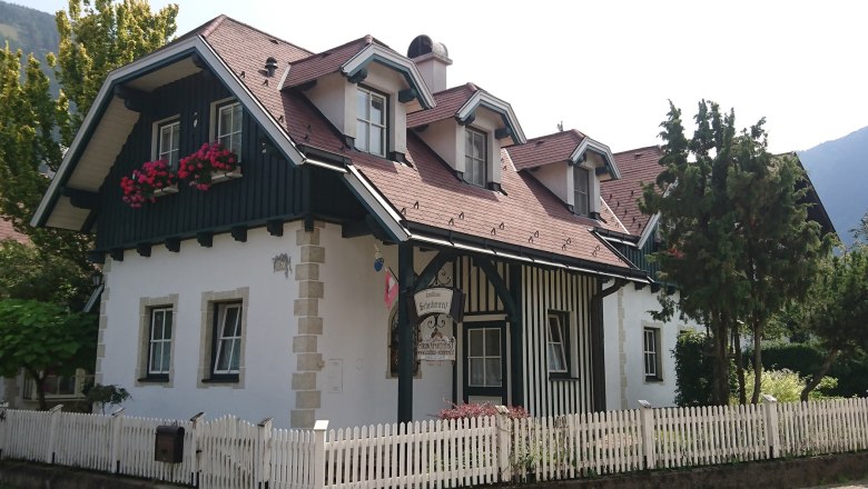 Ein traditionelles Landhaus mit Holzverkleidung, roten Dachziegeln und Blumenk&auml;sten vor den Fenstern. Ein wei&szlig;er Holzzaun umgibt das Grundst&uuml;ck.