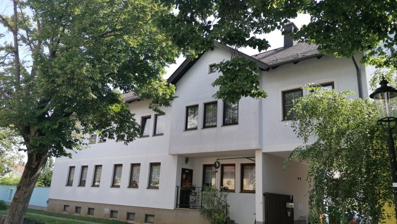 Ein weißes Gebäude mit mehreren Fenstern, umgeben von Bäumen und einem Laternenpfahl.