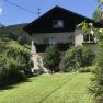 Ein zweistöckiges Haus mit Balkon, umgeben von einem gepflegten Garten und grünen Hügeln im Hintergrund.