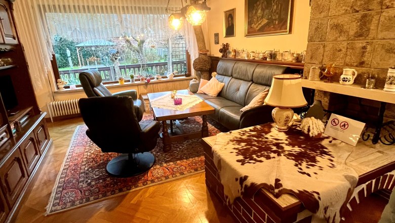 Gemütliches Wohnzimmer mit Ledersofa, Sessel, Holztisch und Kamin. Große Fensterfront mit Blick nach draußen. Rustikale Einrichtung mit Teppich.