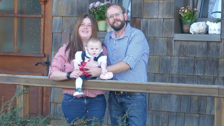 Ein Paar mit einem Baby steht vor einem Holzhaus.