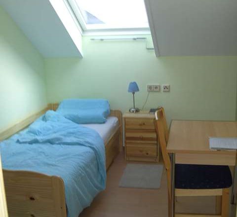 Ein kleines Einzelzimmer mit Bett, Nachttisch, Schreibtisch und Stuhl unter einem Dachfenster.