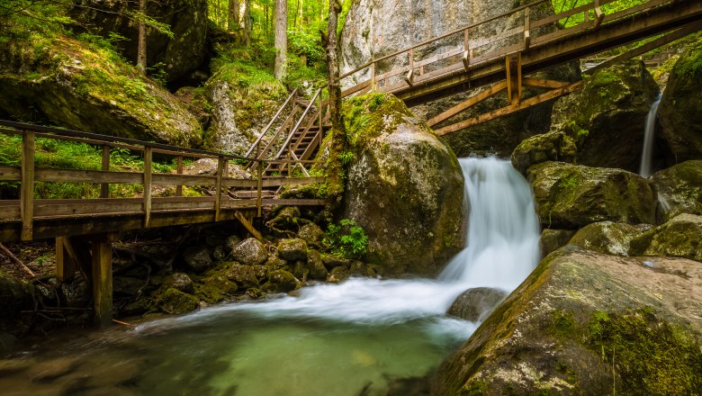 Wasserfall mit Holzbrücken und Treppen in einem bewaldeten Gebiet.