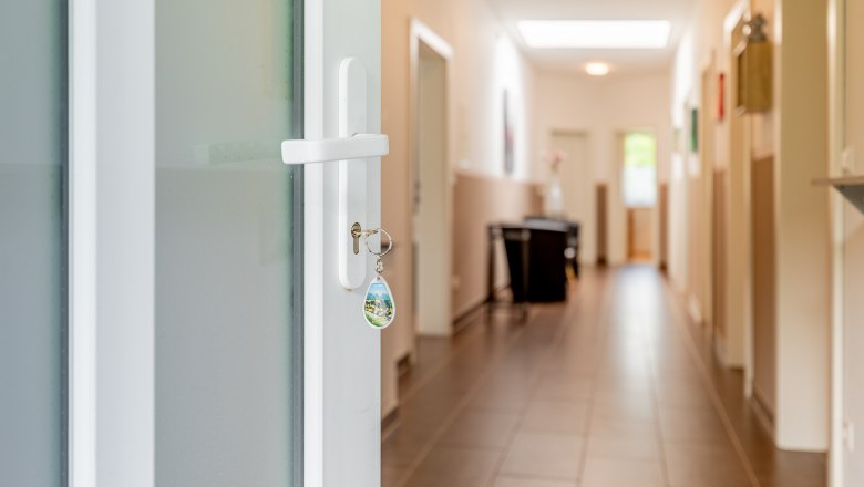 Ein Schl&uuml;ssel steckt in einer wei&szlig;en T&uuml;r, die zu einem hellen Flur mit Fliesenboden f&uuml;hrt.